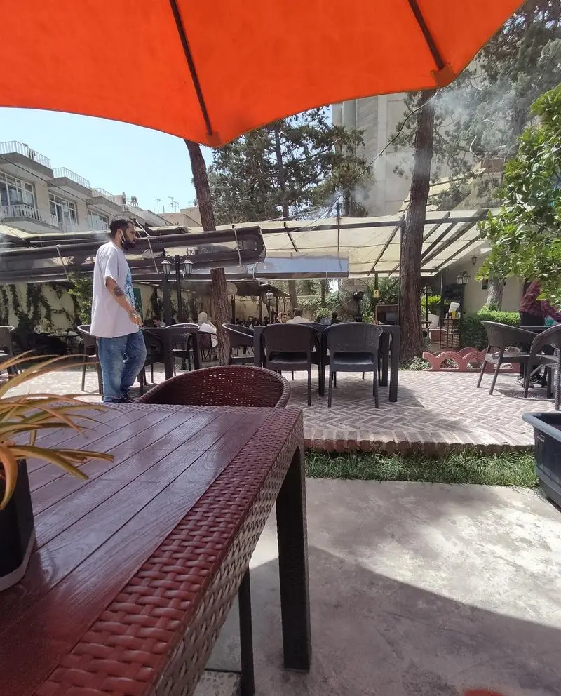 کافه هومان: فضایی دلنشین و باز در روز تابستانی، با مشتریان در حال استراحت زیر چتر نارنجی و میزهای حصیری زیبا.