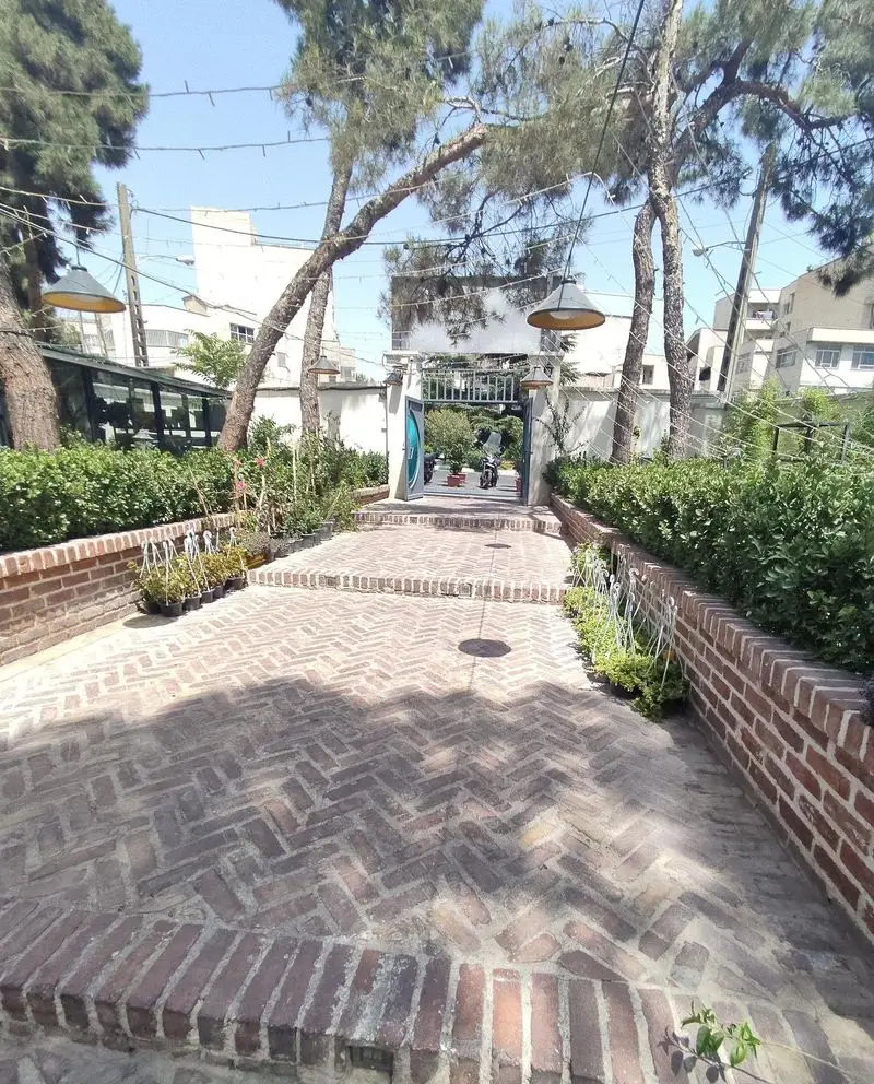 فضای باز دلنشین کافه هومان با مسیر آجری طرح جناغی، درختان سرسبز و نورپردازی زیبا، مکانی آرام برای لذت قهوه.