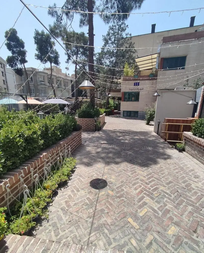 مسیر ورودی دلنشین کافه هومان با سنگفرش آجری، فضای سرسبز و نورپردازی زیبا، محیطی آرام برای کافه گردی در تهران.