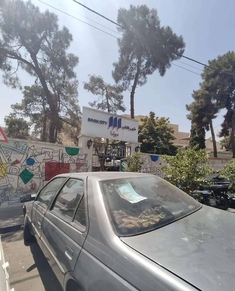 نمای بیرونی کافه هومان در شهر کتاب مینا، ورودی با درختان سرسبز و دیوار نقاشی‌شده. محیطی آرام و دلنشین برای علاقه‌مندان به کتاب و قهوه.