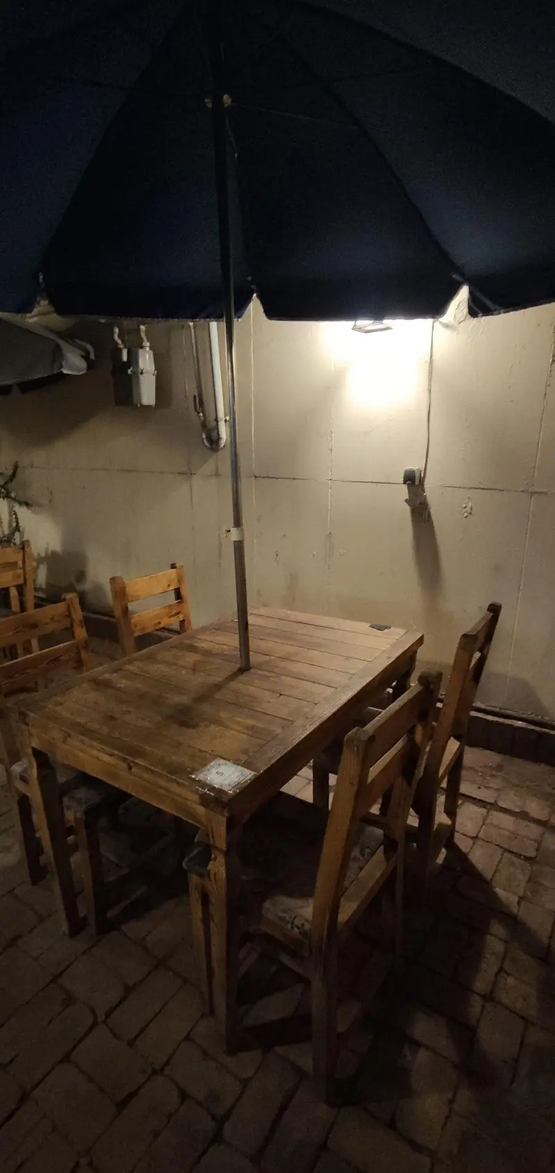 فضای باز و دنج کافه حوض کاشی: میز و صندلی‌های چوبی روستیک با چتر بزرگ آبی، کف آجری و نورپردازی دلنشین برای مشتریان.