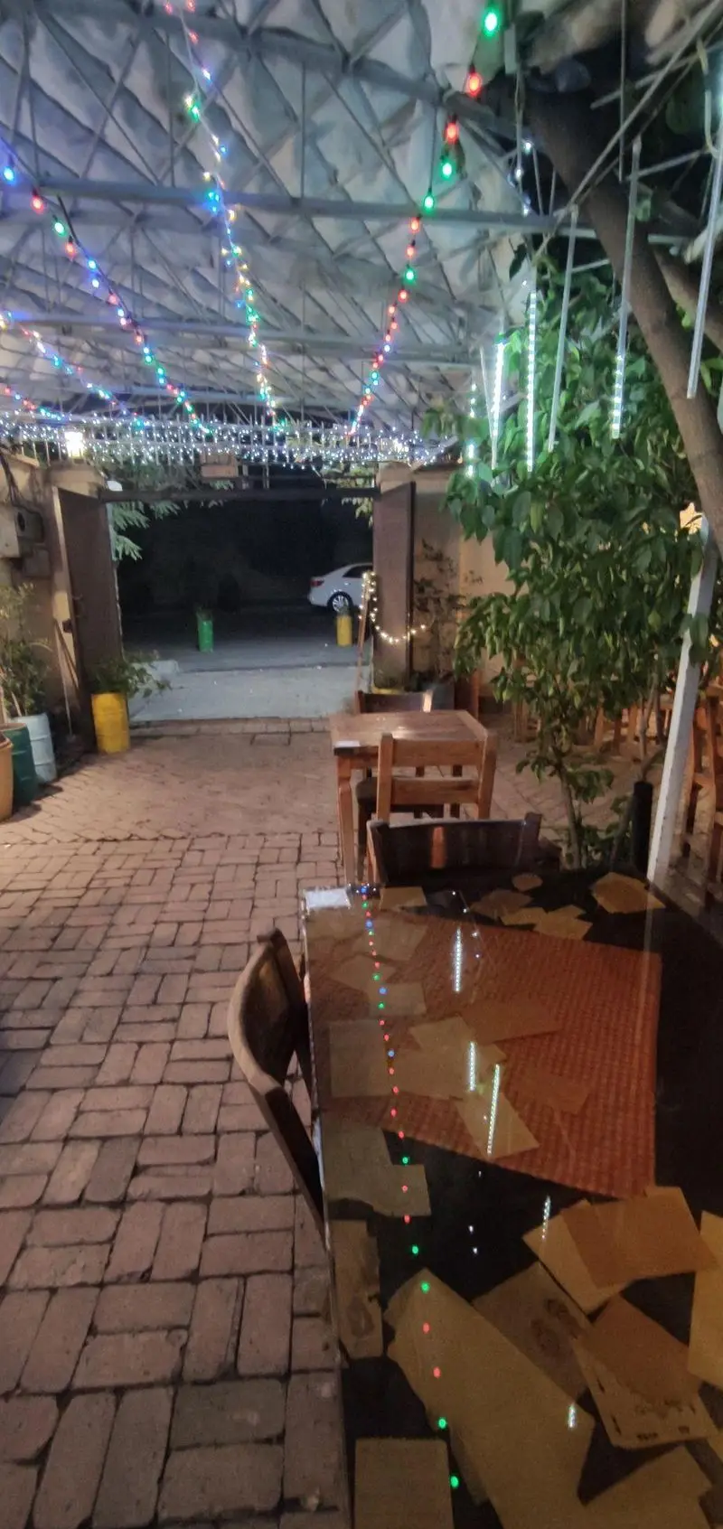 فضای بیرونی دنج و زیبای کافه حوض کاشی در شب با سقف پوشیده از چراغ‌های رنگی، میز و صندلی‌های چوبی و کفپوش آجری، آماده پذیرایی.