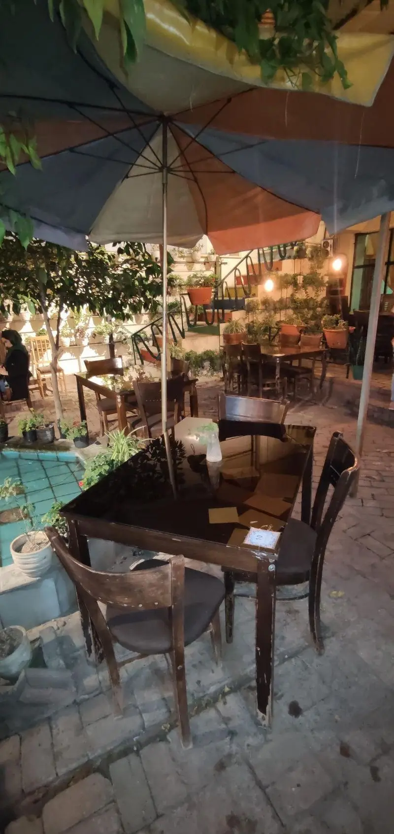 فضای دلنشین و سرسبز کافه حوض کاشی در شب با میز و صندلی‌های چوبی، حوض کاشی‌کاری و چتر بزرگ رنگی، محیطی آرام را برای مشتریان فراهم می‌کند.