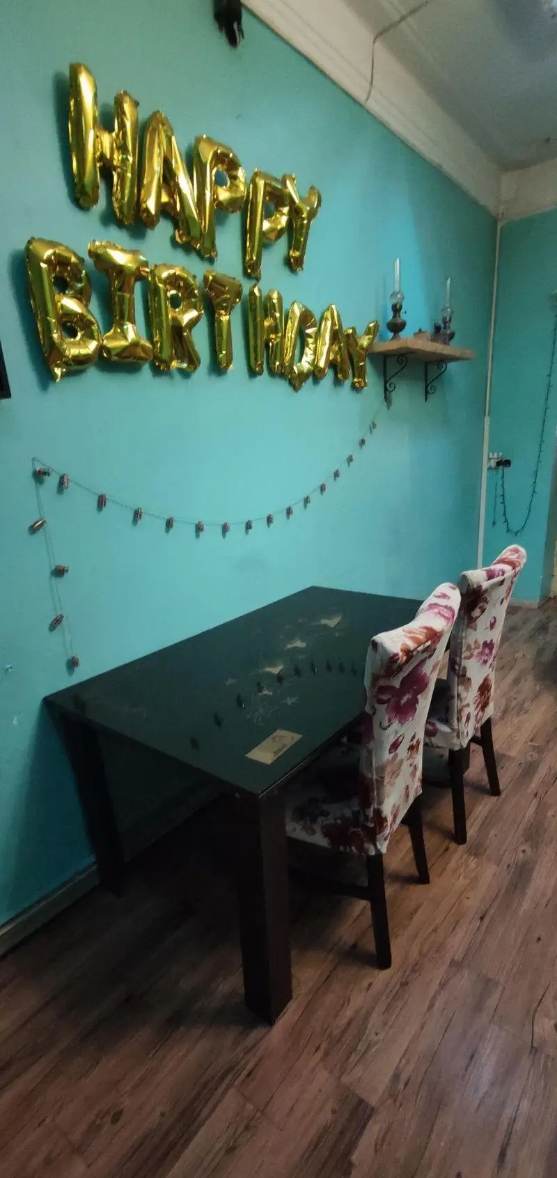 تصویری از فضای داخلی کافه حوض کاشی با دیوار فیروزه‌ای، بادکنک‌های "هپی بیرثدی"، میز و صندلی‌های شیک برای دورهمی و جشن تولد.