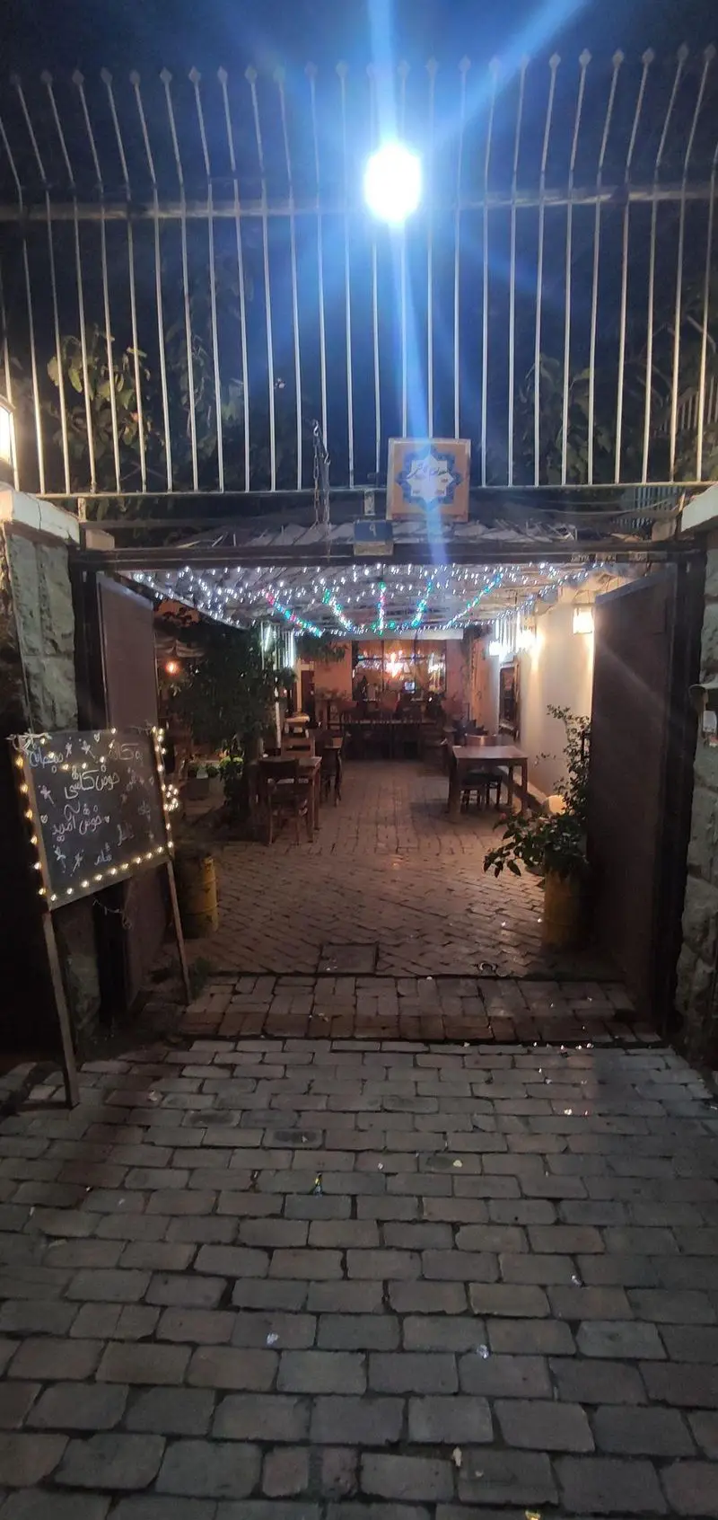 ورودی زیبا و نورانی کافه حوض کاشی در شب، با ریسه های رنگی، فضای نشیمن باز، تابلوی خوش آمدگویی و مسیر سنگفرش.