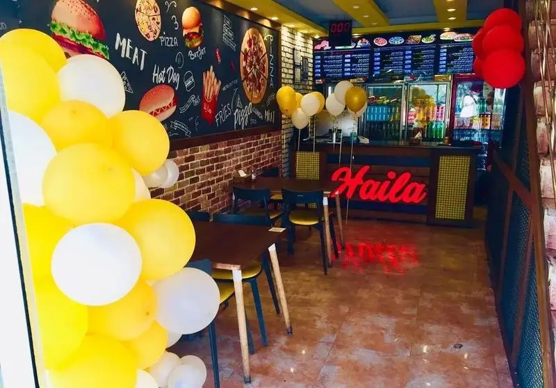 دکوراسیون داخلی رستوران فست فود هایلا با دیوار گچی جذاب، میز و صندلی مدرن، تابلو نئون Hailia و منوی غذاهای متنوع پیتزا، برگر و هات داگ.