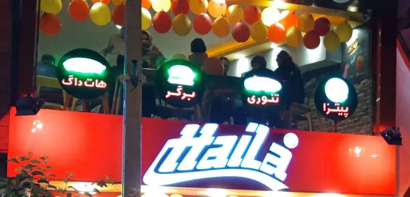 تابلوی فست فود هایلا در شب، با نمایش گزینه‌های غذایی مانند هات داگ، برگر، تنوری و پیتزا. فضای داخلی با مشتریان و بادکنک‌های رنگی.