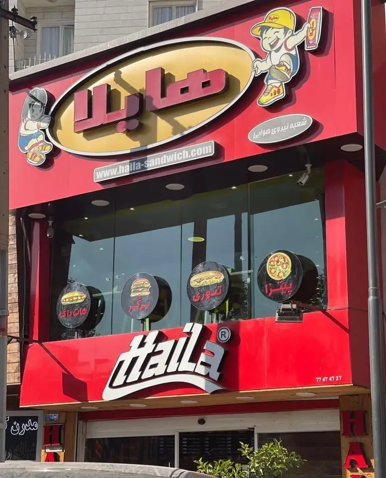 نمای خارجی رستوران ساندویچ هایلا (Hailà) با تابلوی اصلی، شعبه نیروی هوایی و آدرس سایت haila-sandwich.com. منوی پیتزا، برگر و هات داگ.