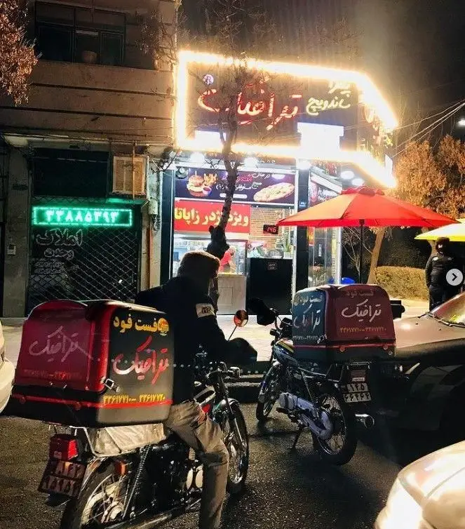 نمای شبانه رستوران فست فود ترافیک با تابلوی نئون روشن و موتورسیکلت‌های پیک آماده تحویل سفارشات آنلاین غذا.