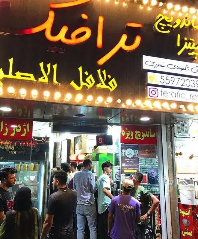 نمای شلوغ از فست فود ترافیک در شب با نورپردازی نئونی، مشتریانی که برای ساندویچ، فلافل و پیتزای اصلی انتظار می‌کشند.