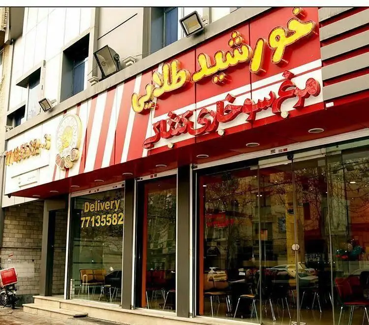 نمای بیرونی فست فود خورشید طلایی، با تابلوی مشخص کننده مرغ سوخاری و شاورما، به همراه شماره تماس دلیوری روی شیشه.