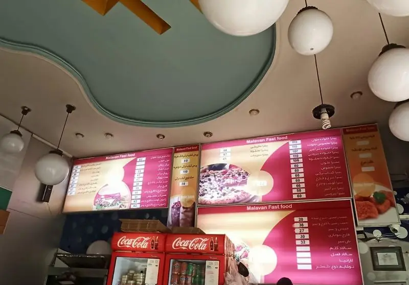 تصویری از داخل فست فود ملوان با تابلوهای منوی غذا، پیتزا، ساندویچ و نوشیدنی به همراه یخچال‌های کوکاکولا.