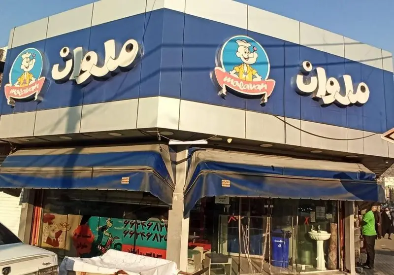 نمای بیرونی فست فود ملوان با تابلوی درخشان آبی و نقره‌ای، لوگوی جذاب ملوان زبل و سایه‌بان‌های آبی جلوی مغازه در خیابان.