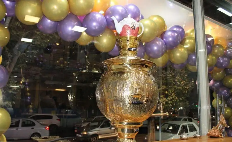 سماور طلایی باشکوه و قوری سفید در ویترین باقلوا فروشی علی کیپا پیروزی، مزین به بادکنک‌های طلایی و بنفش در جشن افتتاحیه.
