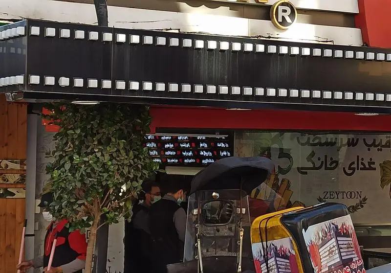 نمای بیرونی رستوران فست فود زیتون با تابلوی طرح فیلم سینمایی، نشان‌دهنده غذای خانگی و فست فود در فضایی شلوغ شهری.