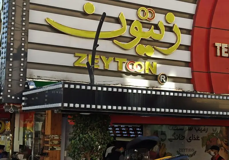 نمای نزدیک از تابلوی رستوران فست فود زیتون (ZEYTOON) با حروف برجسته طلایی و طراحی نوار فیلم، نشان دهنده برند و خدمات غذایی.