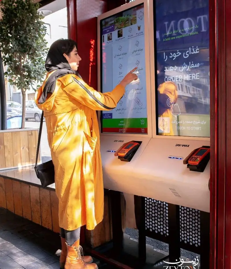 مشتری در حال استفاده از کیوسک سفارش‌گیری سلف سرویس رستوران فست فود زیتون برای سفارش غذا، تجربه خرید آسان و مدرن.
