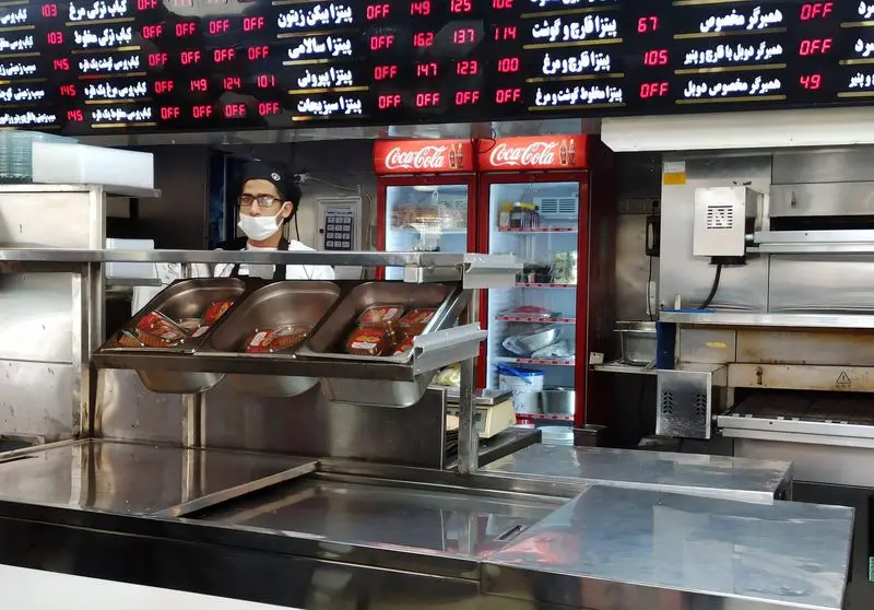 نمایی از آشپزخانه رستوران فست فود زیتون با سرآشپزی پشت پیشخوان استیل، یخچال‌های کوکاکولا و منوی دیجیتال پیتزا، کباب و همبرگر.