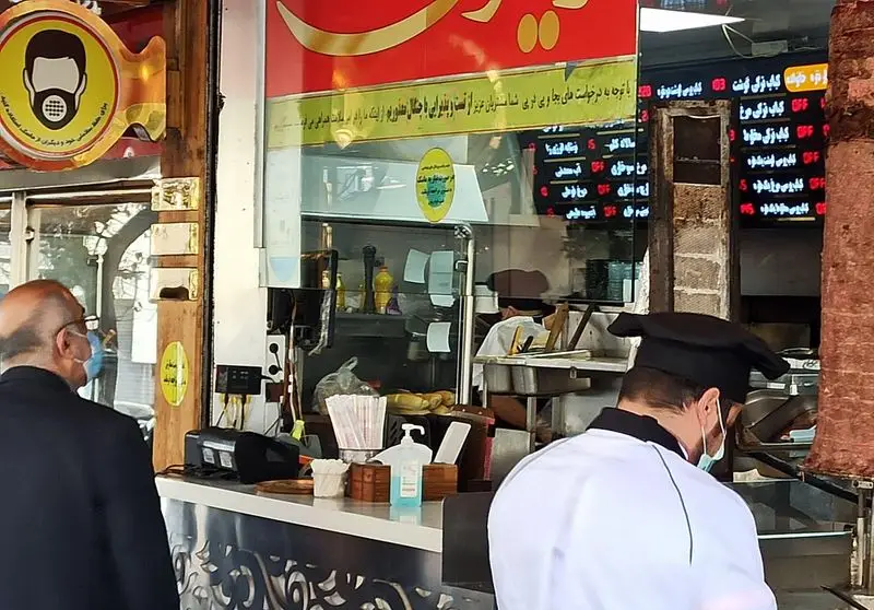 رستوران فست فود زیتون، با نمایی از آشپز در حال آماده‌سازی کباب ترکی و مشتری ماسک‌زده کنار میز سفارش، تاکید بر رعایت بهداشت.
