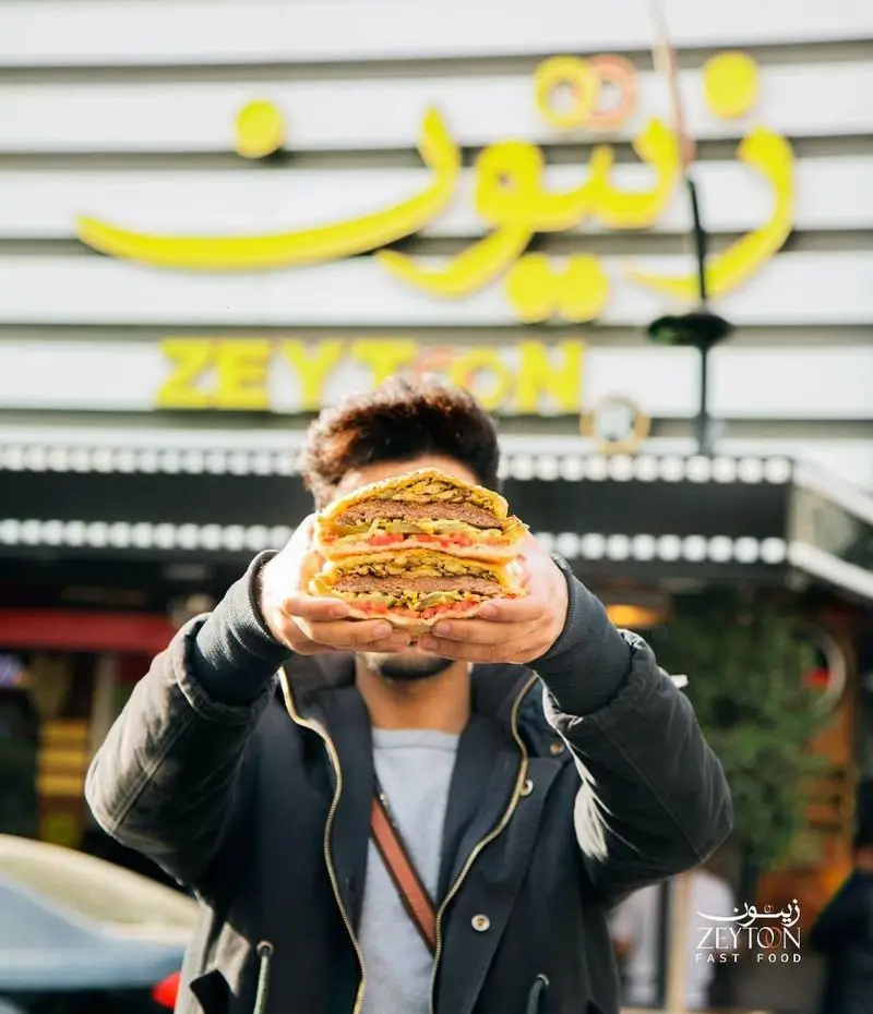 برگر بزرگ و آبدار فست فود زیتون در دست مشتری، با تابلوی رستوران زیتون در پس زمینه، نمادی از کیفیت و طعم.