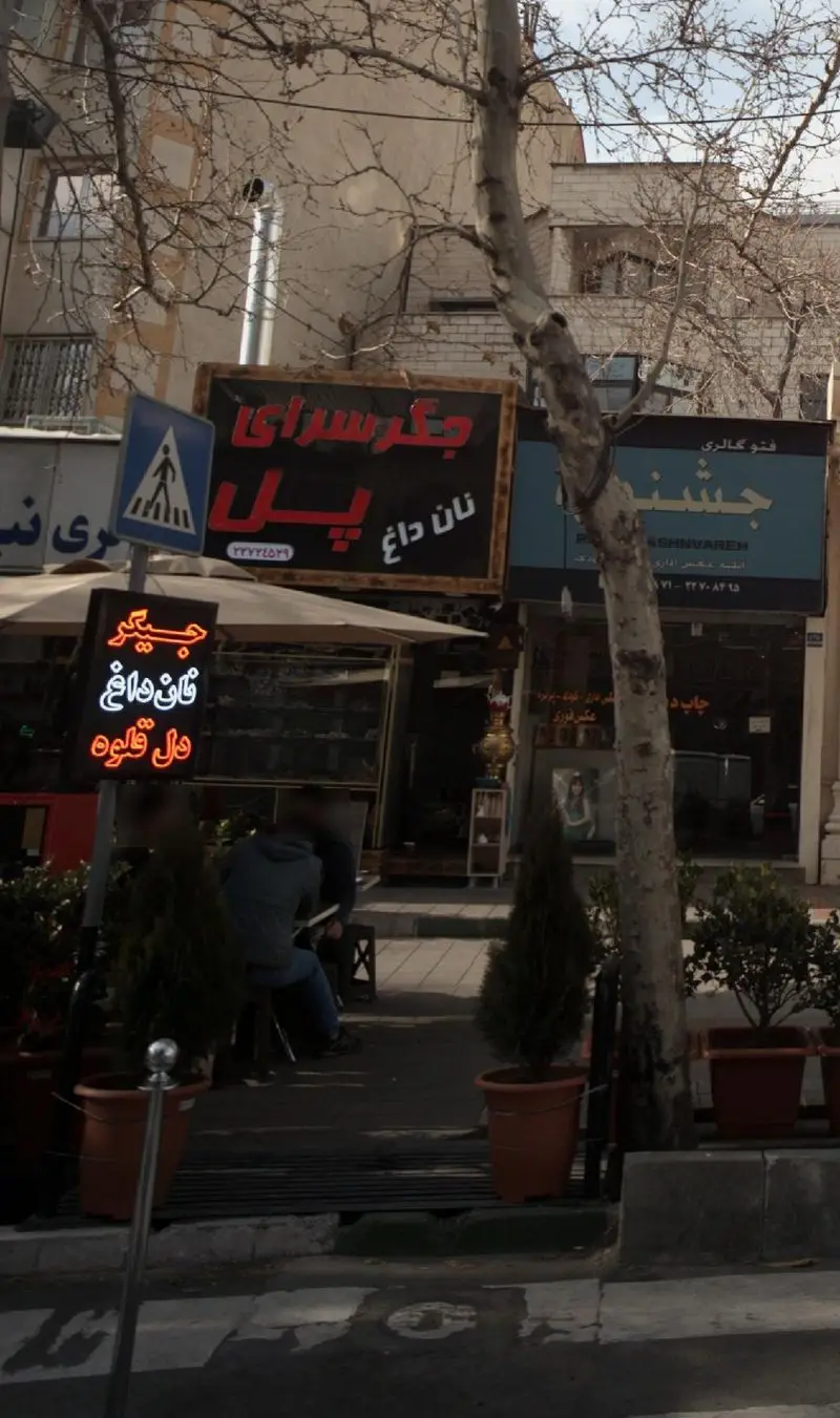 تصویری از نمای بیرونی جگر سرای پل، با تابلوهای نان داغ و دل و قلوه. مردم در حال استراحت در فضای باز، در یک روز روشن شهری.