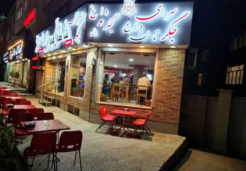 نمای بیرونی جگرکی کبابی شاطر میرزا در شب با تابلو نورانی، میز و صندلی‌های قرمز در فضای باز و داخل رستوران با مشتریان.