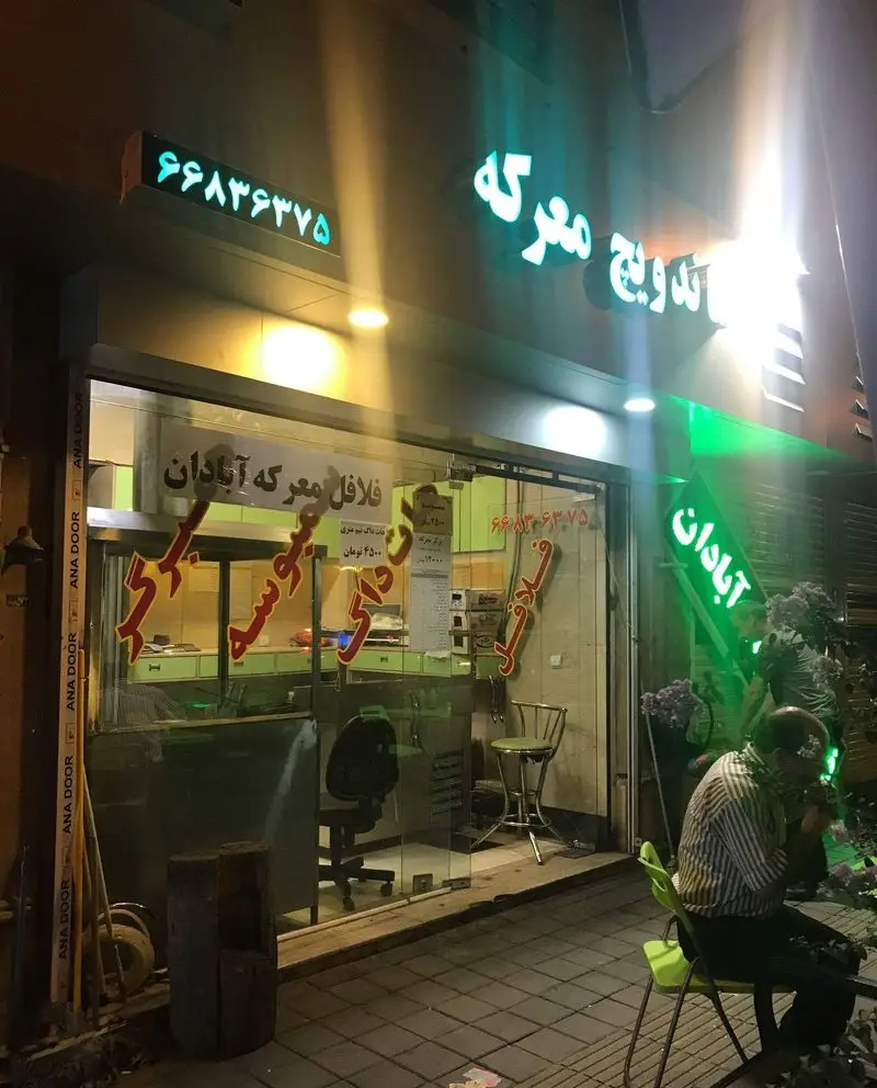 نمای شبانه ساندویچی معرکه آبادان با تابلوهای نئونی روشن و شماره تلفن ۶۶۸۳۶۳۷۵، ارائه‌دهنده فلافل و انواع ساندویچ.