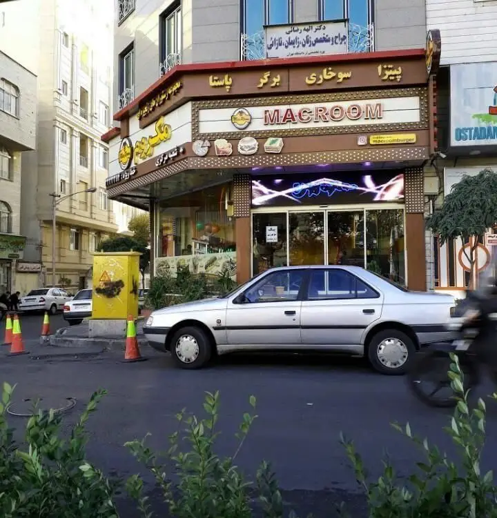 پیتزا مکروم (MACROOM): نمای بیرونی رستوران ارائه دهنده پیتزا، برگر، سوخاری و پاستا در خیابان، با خودروی نقره‌ای جلوی آن.