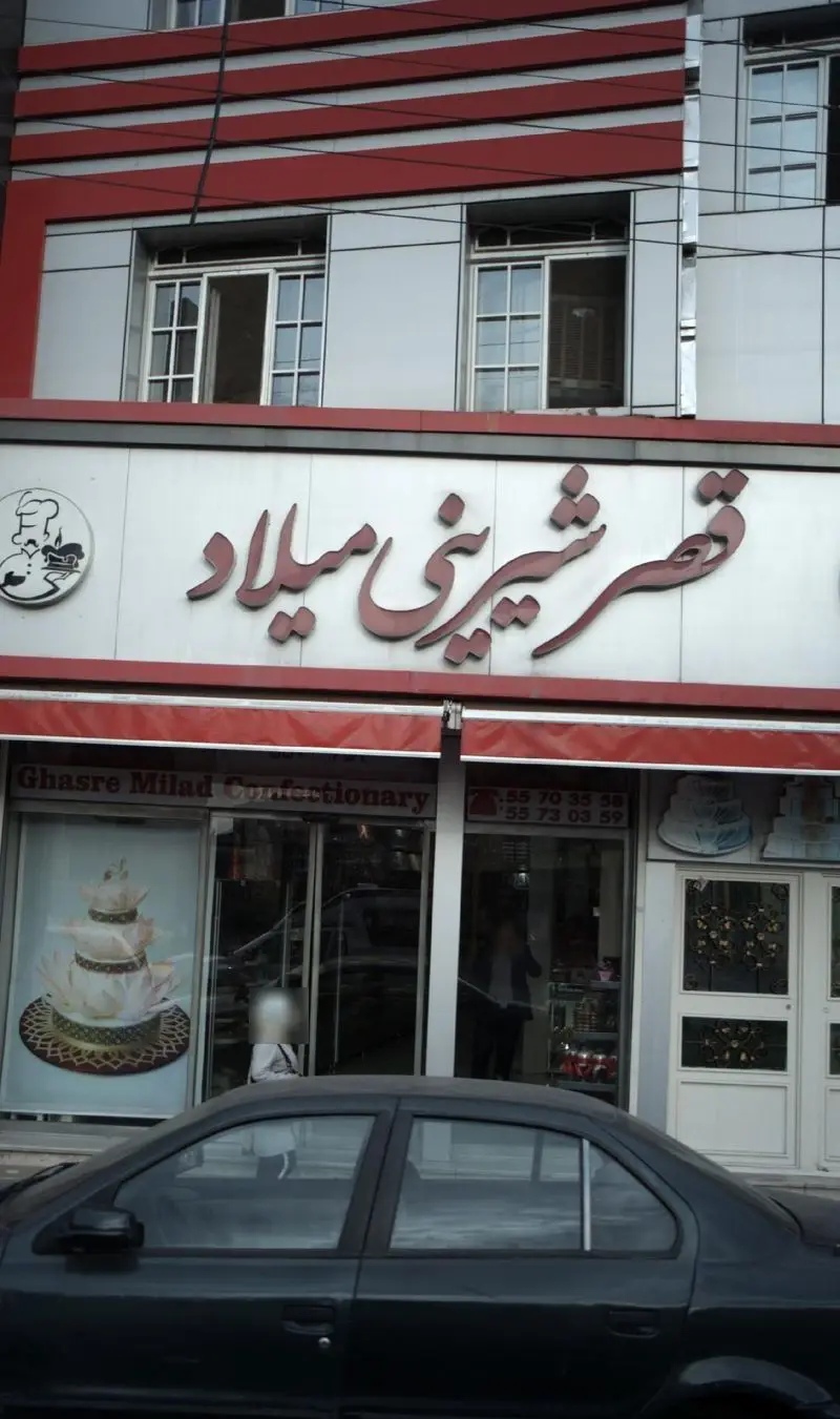 تصویر نمای بیرونی قصر شیرینی میلاد، یک قنادی با تابلوی برجسته فارسی و انگلیسی، ویترین جذاب کیک‌های خاص و شماره تماس.