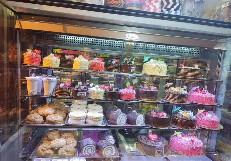 تصویری جذاب از ویترین قنادی شیرینی آنا، شامل انواع کیک‌های تولد، رولت، نان خامه‌ای و دسرهای مدرن و لذیذ. بهترین شیرینی‌ها.