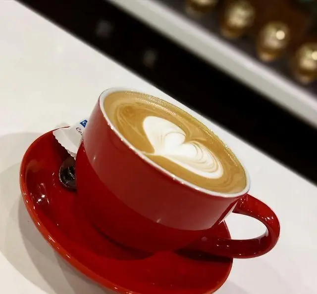 قهوه لاته تازه با طراحی قلبی (لاته آرت) در فنجان قرمز و نعلبکی هماهنگ در کافه سیدمهدی. لحظات آرامش‌بخش را تجربه کنید.