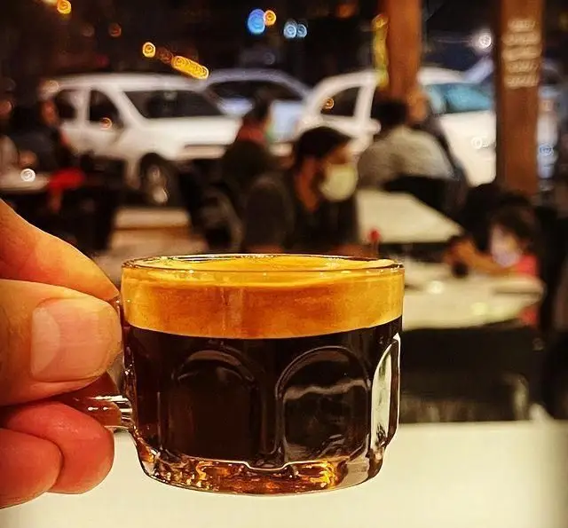 اسپرسو غلیظ با کرمای طلایی در لیوانی کوچک، در دست مشتری کافه سیدمهدی. پس‌زمینه نشان‌دهنده فضای دلنشین شبانه و پر جنب و جوش کافه است.