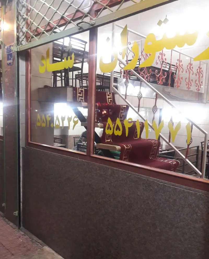 نمایی از ورودی رستوران شاد با درب و پنجره شیشه‌ای، پله‌های داخلی فرش قرمز و میز و صندلی‌ها در طبقات مختلف، با نوشته و شماره تماس روی شیشه.