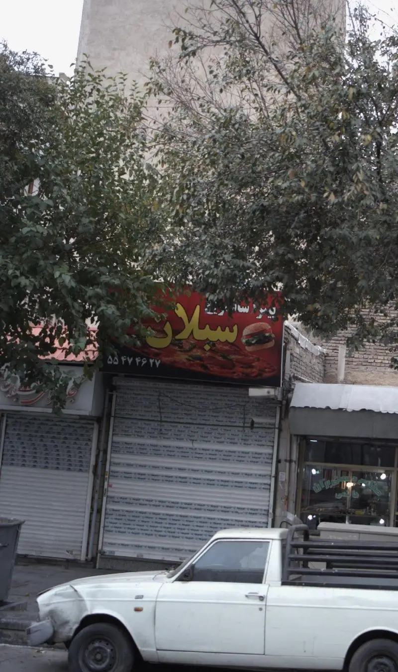 تابلو فست فود پیتزا ساندویچ سبلان شامل عکس پیتزا، همبرگر و شماره ۵۵۴۷۴۶۲۷. مغازه با کرکره‌های بسته و وانت سفید در جلو.