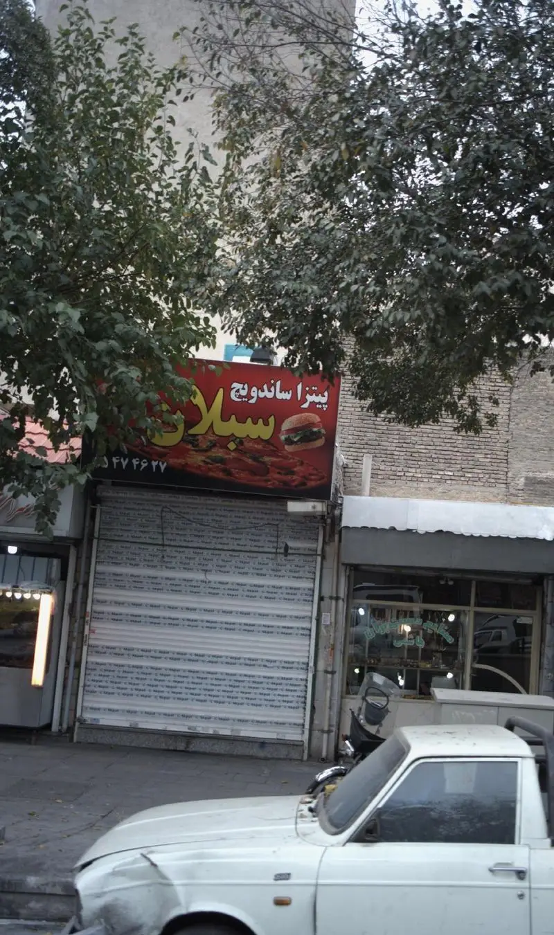 تابلوی فست فود "پیتزا ساندویچ سبلان" با تصویر پیتزا و همبرگر، مغازه بسته شده در خیابان، همراه با درخت و وانت سفید رنگ.