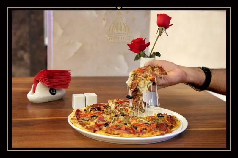 پیتزا کشدار و خوشمزه در مجموعه پذیرایی خیام با پنیر آب شده، همراه با دستمال کاغذی، نمکدان و گل رز قرمز روی میز چوبی.