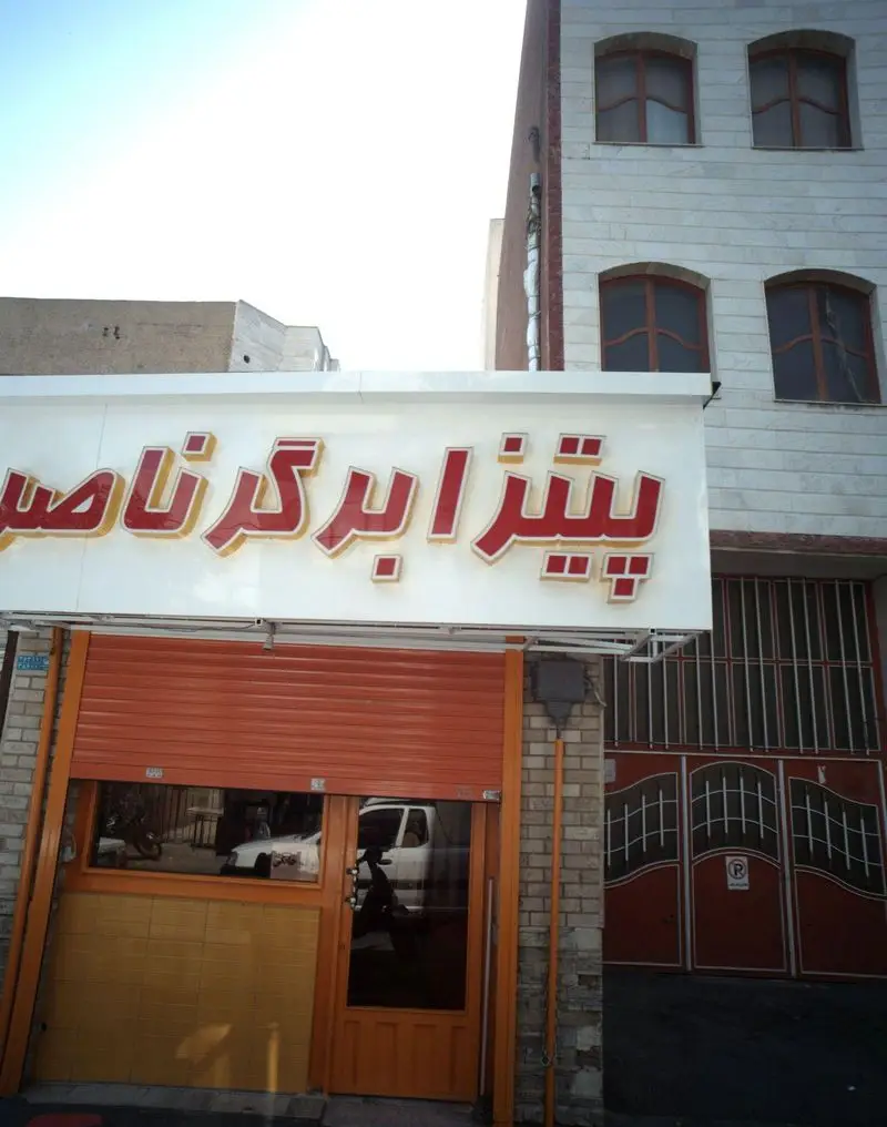 نمای خیابانی و جذاب کسب و کار فست فود پیتزا برگر ناصر با تابلوی روشن، درب کرکره‌ای نارنجی و ساختمان مسکونی بالا