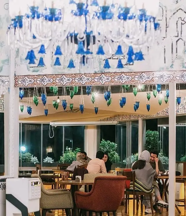 منظره ای از کافه رستوران شمرون با دکوراسیون چشم نواز شامل لوسترهای آبی و آویزهای شیشه‌ای. مشتریان در حال لذت بردن از محیط و غذا.