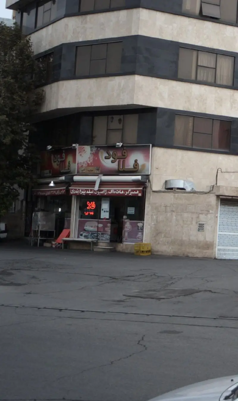 تصویری از نمای بیرونی فست فود فلافود در یک ساختمان، با تابلوی روشن و جزئیات منو شامل همبرگر، پیتزا، هات داگ و ساندویچ.