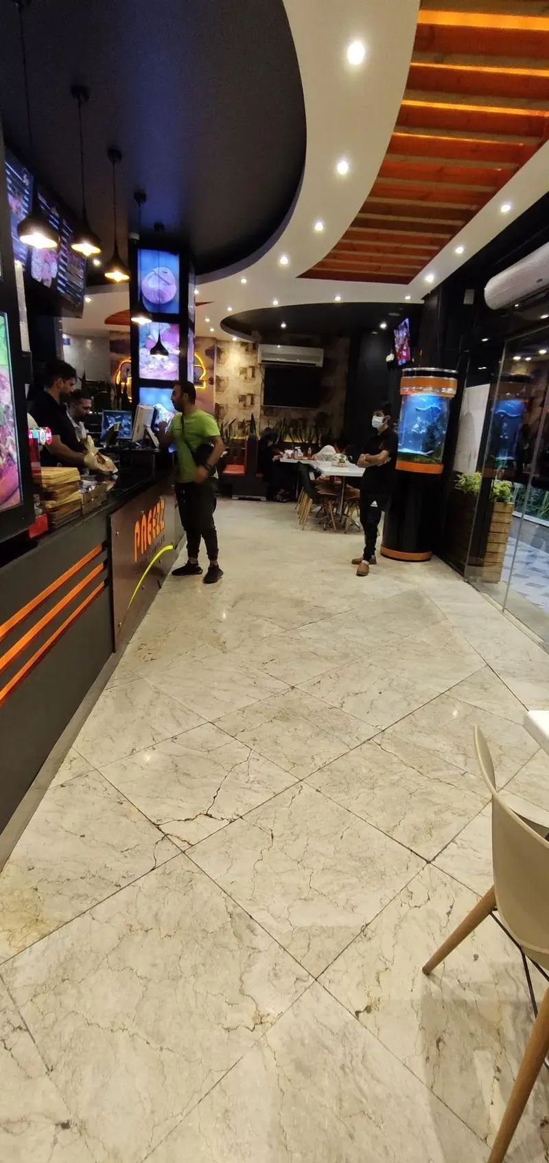 نمای داخلی فست فود پاییز: پیشخوان سرویس شلوغ، کارکنان و مشتریان در محیطی مدرن با نورپردازی زیبا و کفپوش طرح مرمر.