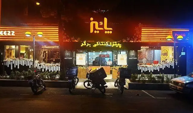 نمایی از فست فود پاییز در شب با نورهای روشن نئون، ارائه دهنده پیتزا، کنتاکی و برگر، با موتورسیکلت‌های دلیوری جلوی مغازه در تهران.