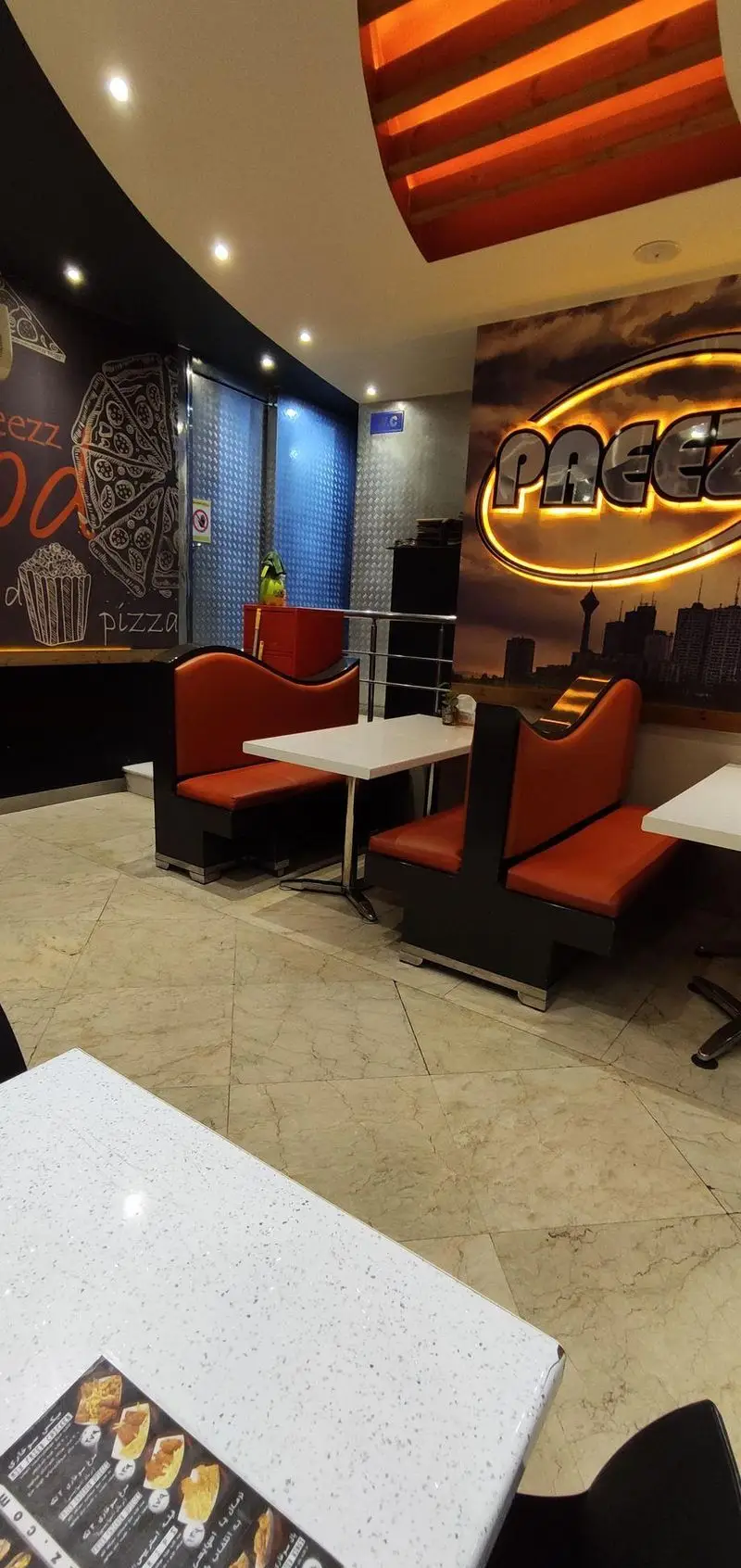 فضای داخلی دنج و مدرن فست فود پاییز با طراحی جذاب، میزهای سفید و صندلی‌های نارنجی-مشکی، لوگوی نورانی و دیوارنگاره پیتزا.