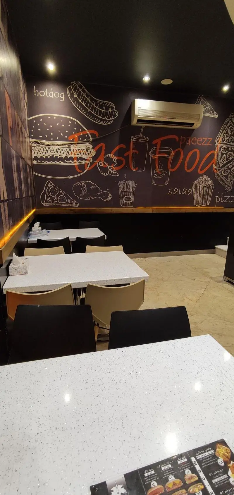 نمای داخلی فست فود پاییز؛ دیوارهای تیره با طرح‌های برجسته برگر، پیتزا، هات داگ و نوشیدنی. میزهای گرانیتی و صندلی‌های مدرن.