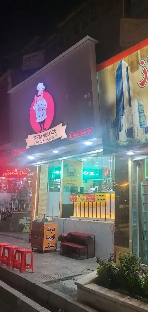 نمای خارجی فست فود ژِ بِ سِ در شب، با تابلوی نئونی Pasta Veloce، ارائه دهنده غذاهای سریع و خوشمزه از جمله پاستا، اسنک و ذرت لوبیا.
