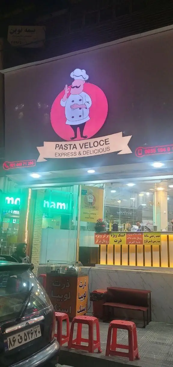 تصویر نمای بیرونی فست فود ژِ بِ سِ با تابلوی بزرگ «پاستا ولوچه اکسپرس و دلیشز» و سرآشپز کارتونی، ارائه دهنده منوی متنوعی از پاستا، برگر و هات داگ.