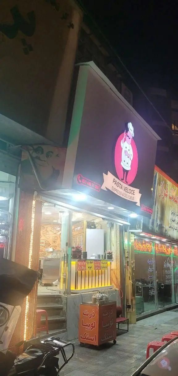 تصویری از نمای شب فست فود ژِ بِ سِ در تهران، با تابلوی Pasta Veloce و ارائه انواع اسنک، ذرت، لوبیا و پاستاهای خوشمزه.