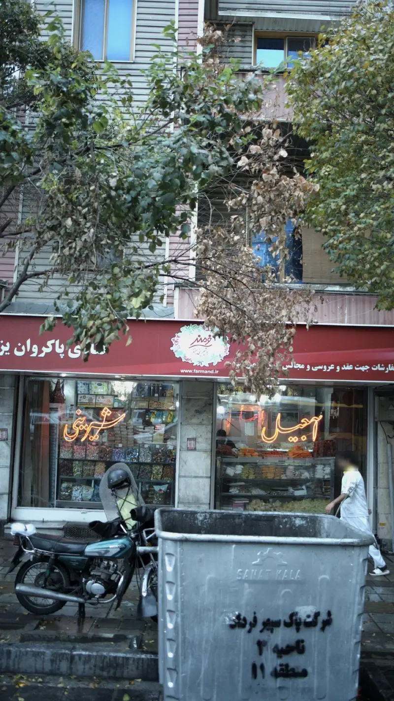 تصویری از نمای بیرونی قنادی پاکروان یزدی (فرماند) با ویترین پر از شیرینی و آجیل، موتور سیکلت و سطل زباله در پیاده‌رو.