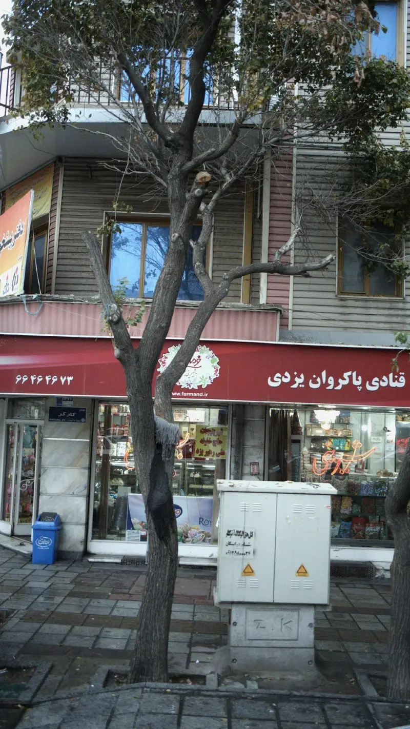 تصویر نمای بیرونی قنادی پاکروان یزدی با تابلوی قرمز، شماره تلفن و لوگوی فرمند، ویترین پر از شیرینی و شکلات، درخت و پیاده‌رو.