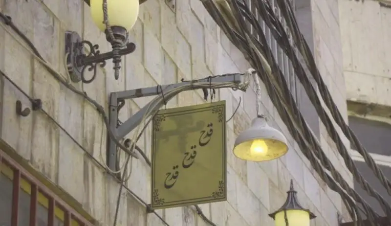 تابلوی رستوران ایرانی قدح با خط نستعلیق روی دیوار سنگی، همراه با چراغ خیابانی و کابل‌های برق آویزان در شهر.