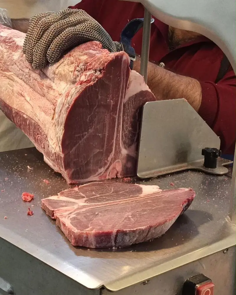 قصاب ماهر در سوپر پروتئین ملل در حال برش گوشت تازه گوساله با دستگاه صنعتی برای تهیه استیک تی بون باکیفیت و تازه.